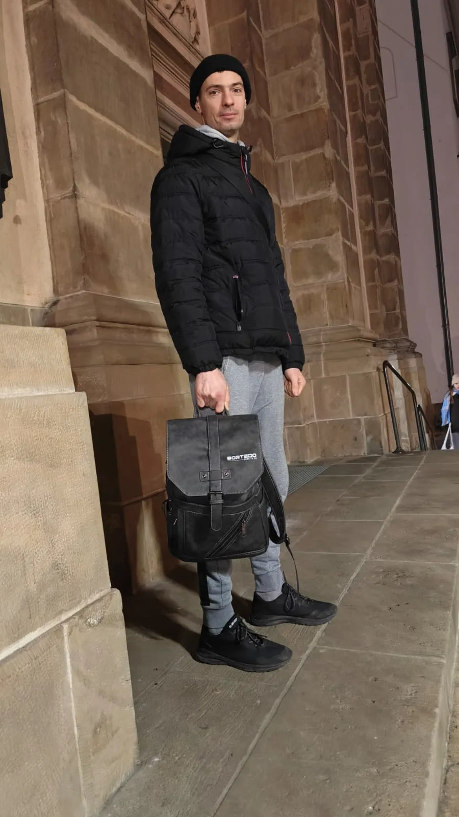 Person in black puffer jacket and gray pants holding a black backpack against a stone building.