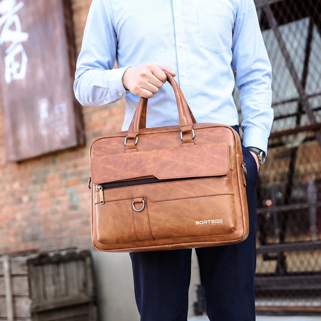 A man in a light blue dress shirt and navy trousers holds a brown textured leather briefcase bag with the brand name BORTEGO printed on the front, set against an urban brick wall background.