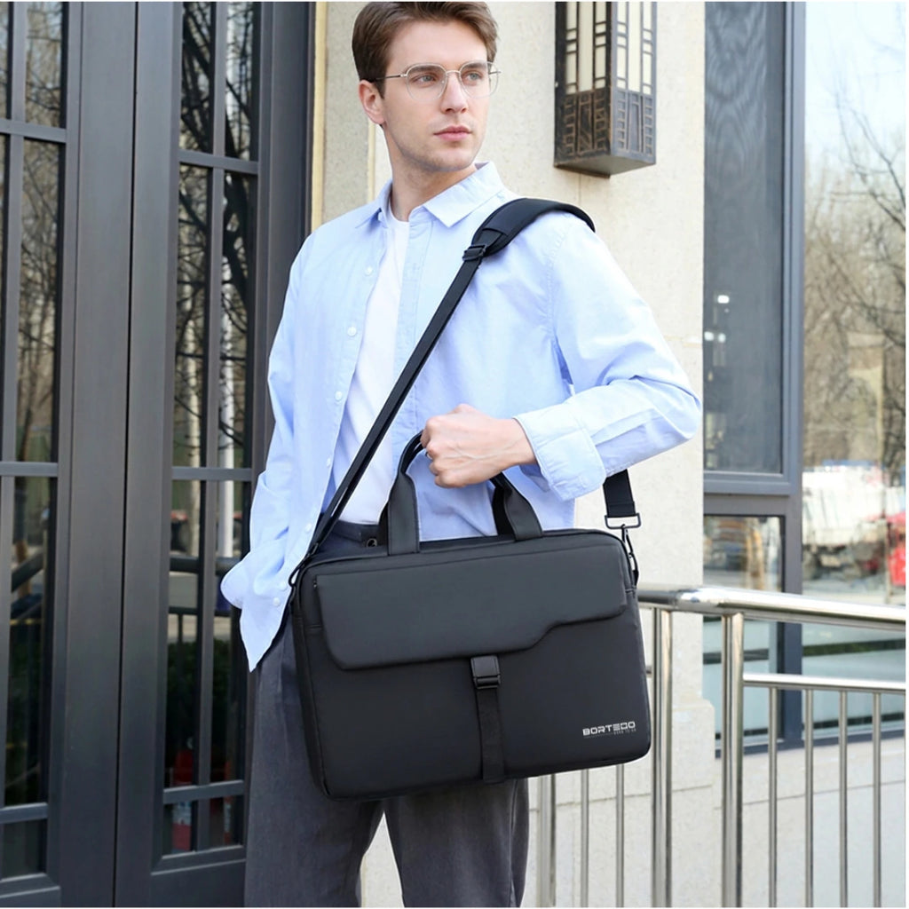 A man wearing a light blue shirt and glasses carries a black briefcase-style shoulder bag with a top handle and adjustable strap, standing outdoors near a building entrance.