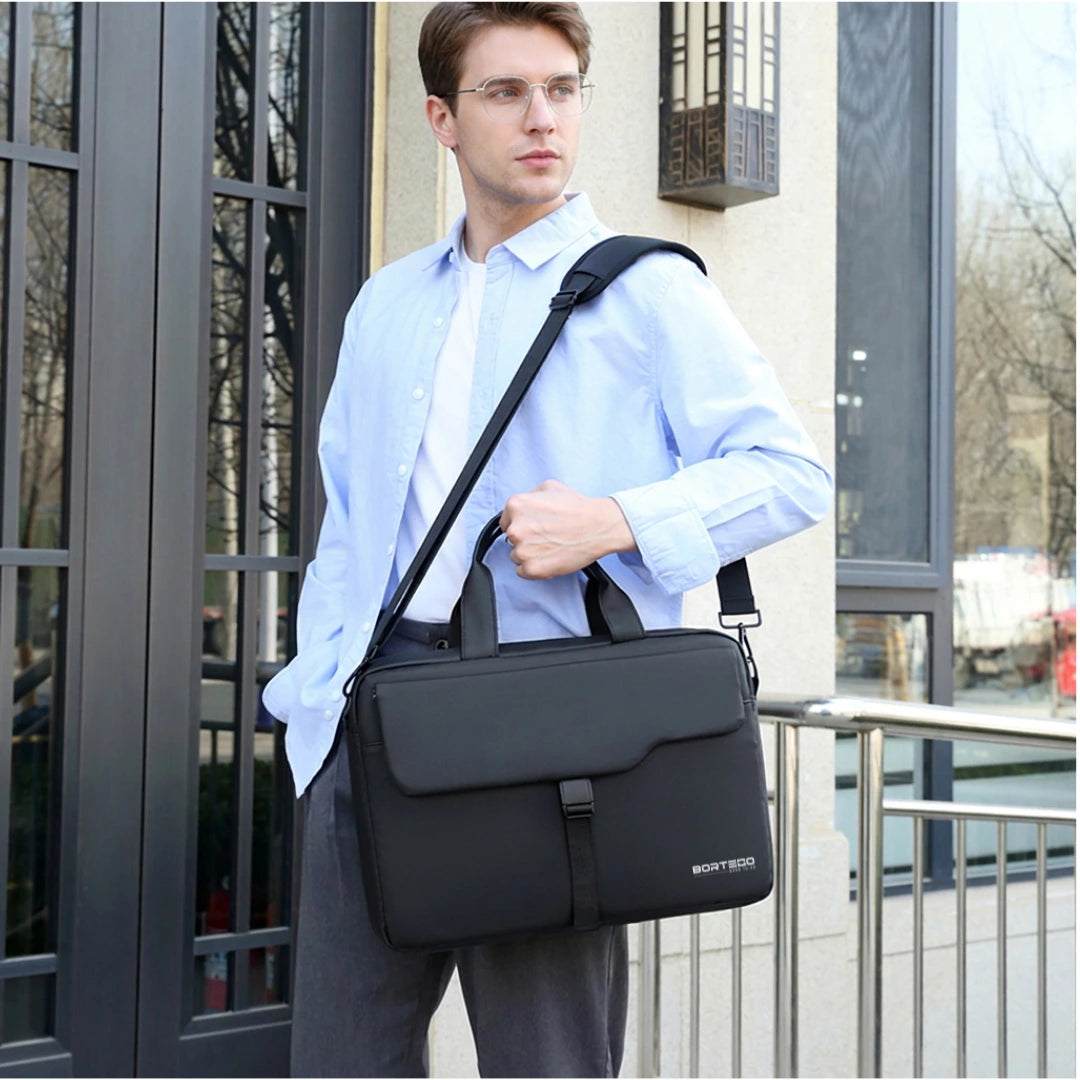 A man wearing a light blue shirt and glasses carries a black briefcase-style shoulder bag with a top handle and adjustable strap, standing outdoors near a building entrance.