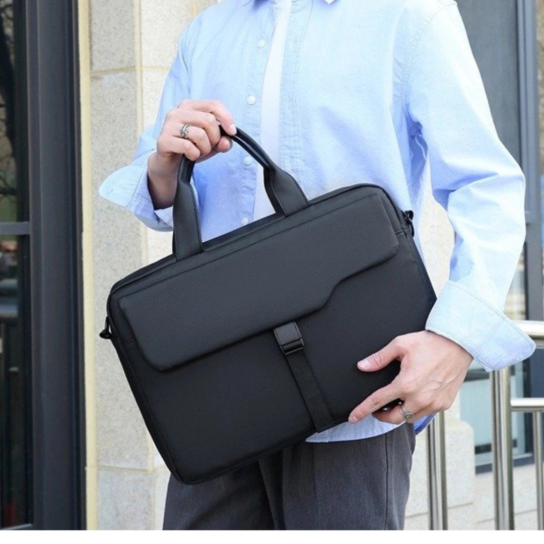 A person in a light blue button-up shirt holds a black nylon briefcase-style bag with top handles and a front flap closure, photographed outdoors near a building entrance.