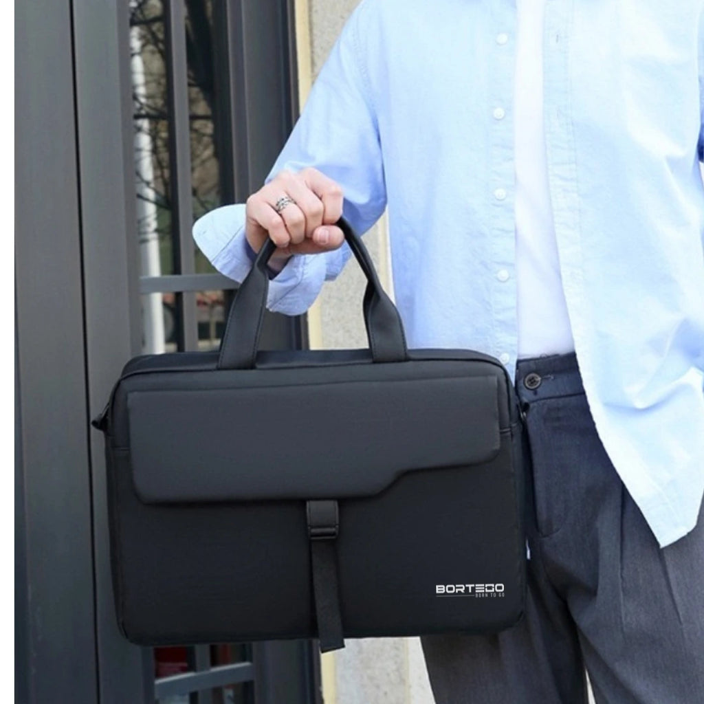 A person in a light blue dress shirt holds a black BORTEGO laptop briefcase bag with a top handle, set against a dark window background.