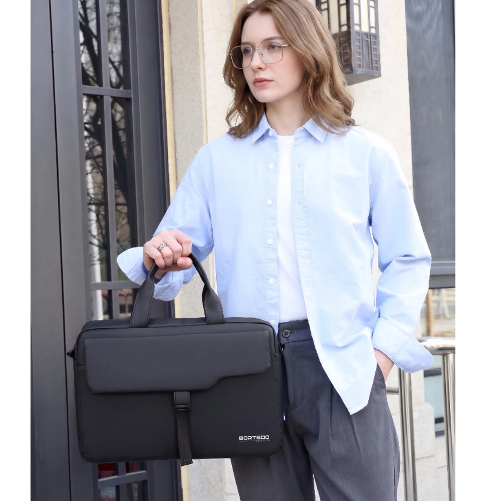 A woman wearing a light blue button-down shirt and gray trousers holds a black briefcase-style bag with a top handle in front of a building entrance.