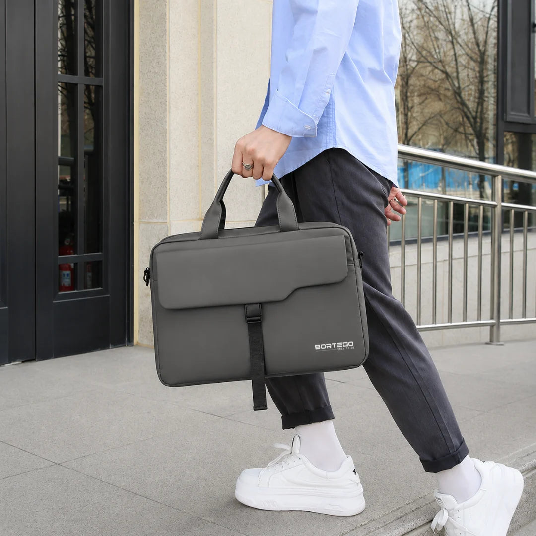 A person in a light blue shirt, dark pants, and white sneakers carries a dark gray laptop bag with a top handle and front strap closure, walking outdoors near a building entrance.