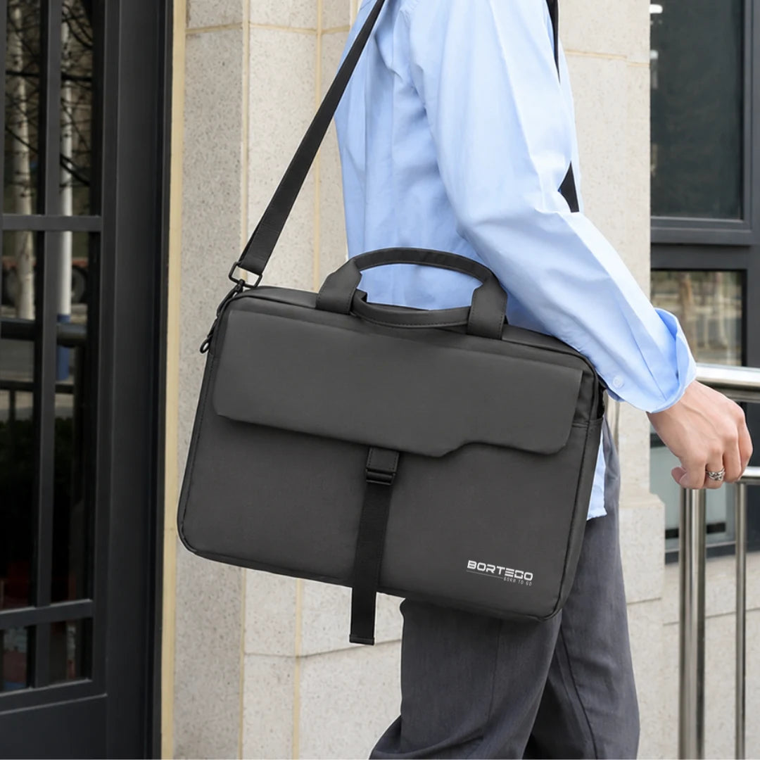 A person in a light blue dress shirt and gray trousers carries a dark gray BORTEGO laptop briefcase bag with a top handle and shoulder strap, standing in front of a stone building.