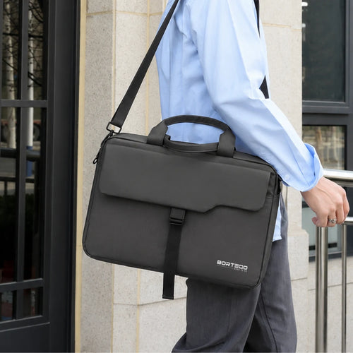 A person in a light blue dress shirt and gray trousers carries a dark gray BORTEGO laptop briefcase bag with a top handle and shoulder strap, standing in front of a stone building.