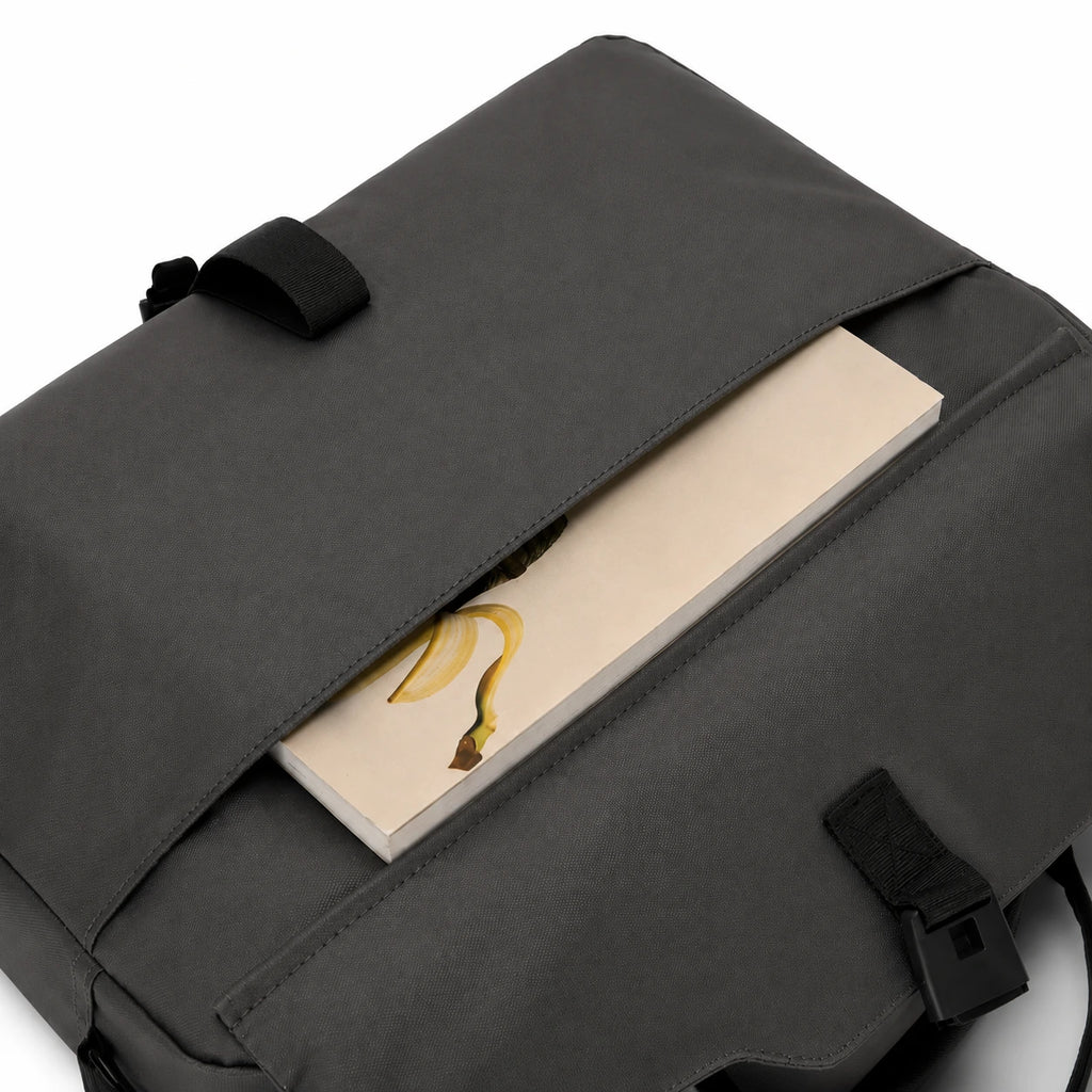 A dark gray backpack shown in a close-up view with a flap pocket open, revealing a notebook with a banana illustration inside. The backpack features black straps and a black plastic buckle closure.