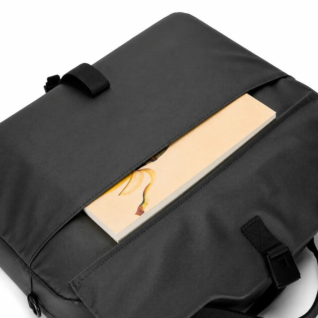 A black nylon messenger bag shown from an angled top-down view, featuring a front flap pocket with a strap closure and a notebook tucked inside.