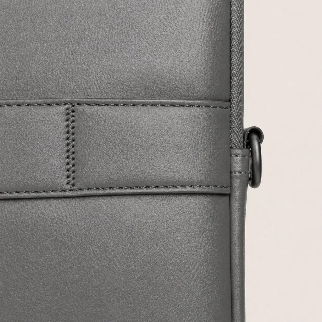 Close-up of a gray textured leather bag showing a stitched panel and a black metal ring detail on a light beige background.