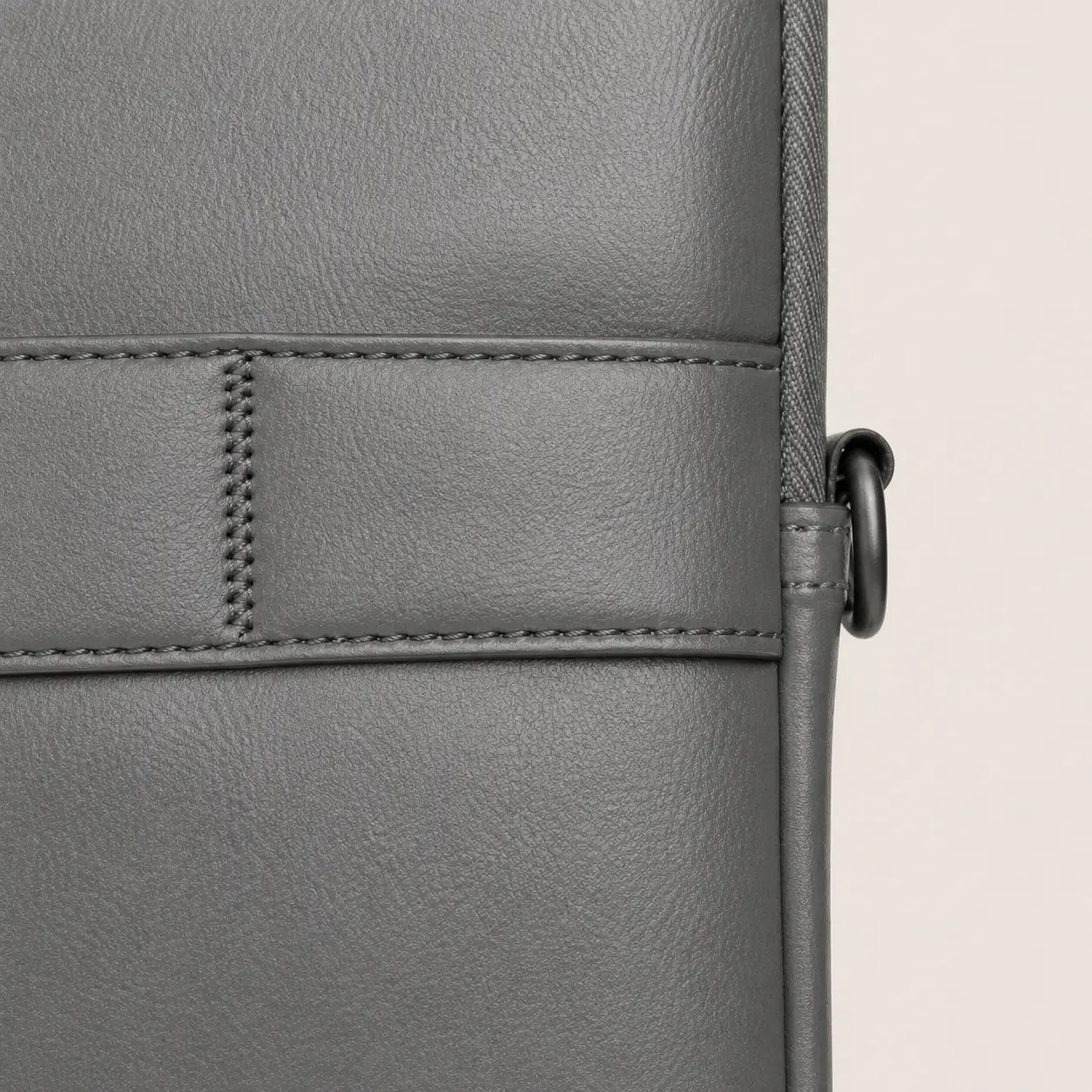 Close-up of a gray textured leather bag showing a stitched panel and a black metal ring detail on a light beige background.