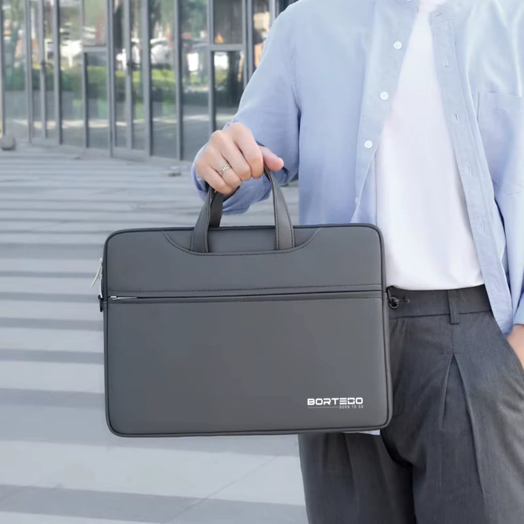 A man in a light blue button-up shirt and gray trousers holds a dark gray BORTEGO laptop sleeve with top handles in an outdoor urban setting.