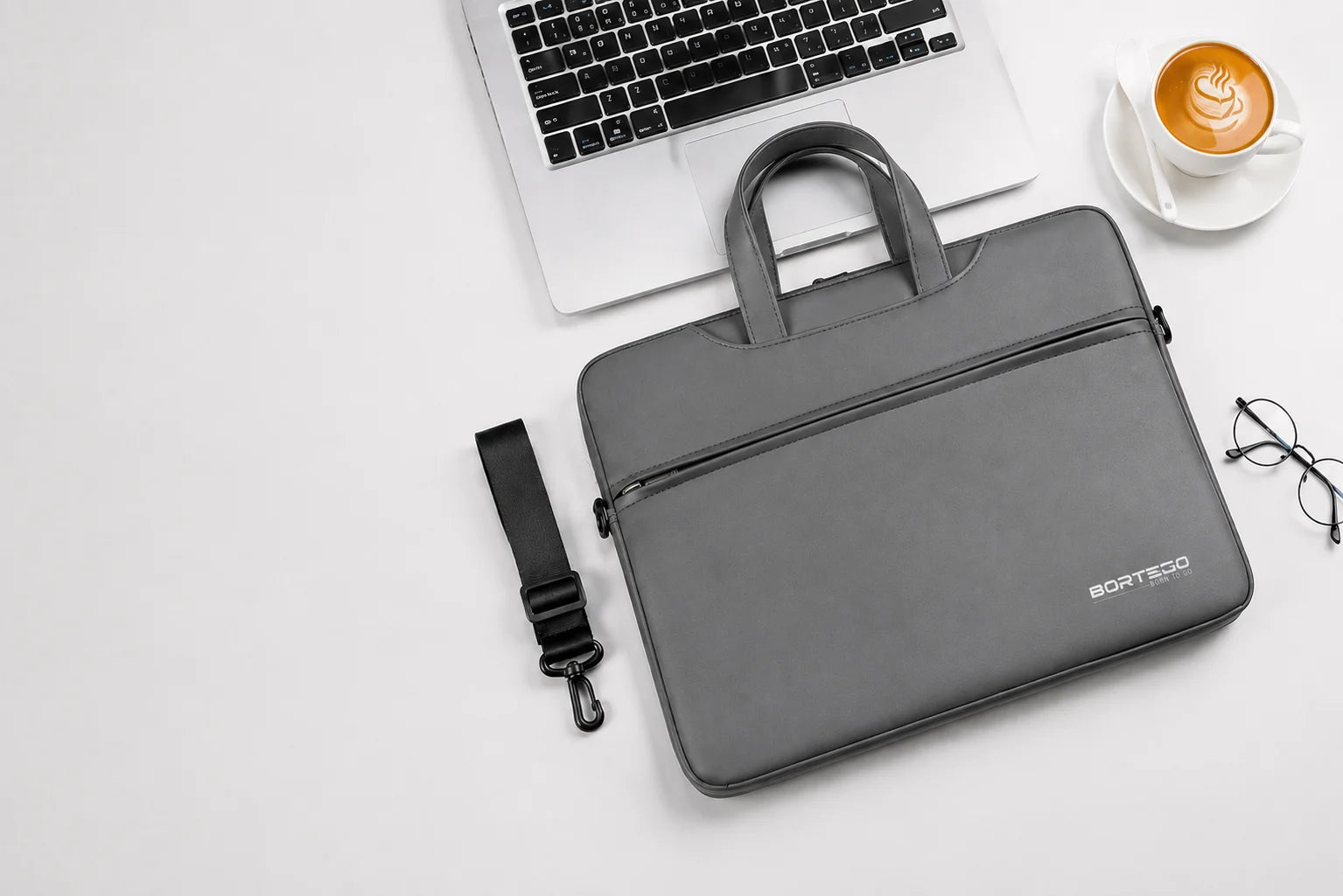 A dark gray BORTEGO laptop sleeve with dual top handles and a detachable shoulder strap, styled in a flat-lay arrangement on a white surface alongside a laptop, a cup of coffee, and eyeglasses.