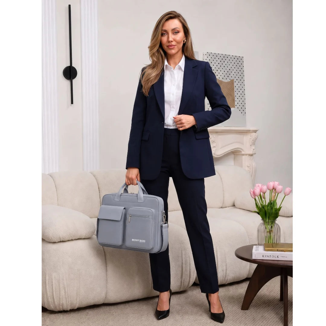 A woman stands in a living room setting wearing a navy blue blazer and matching trousers, paired with a white collared shirt and black heels, while holding a light gray structured briefcase-style bag.