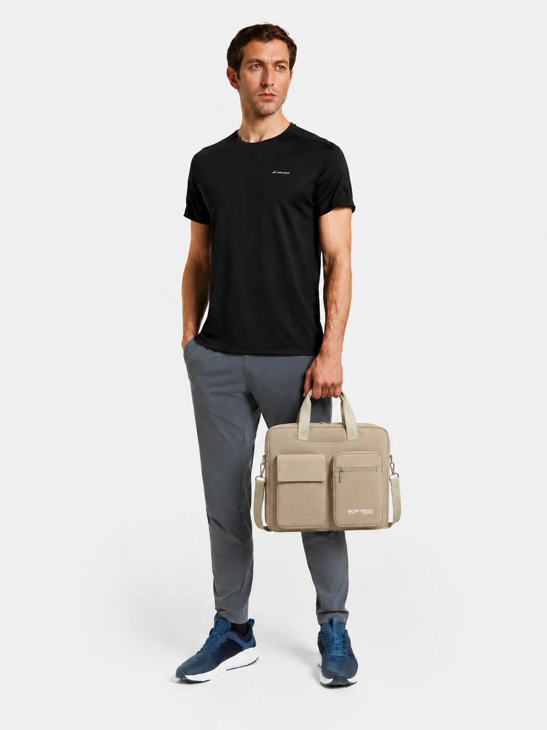 A man wearing a black t-shirt, gray pants, and blue sneakers stands against a light gray studio background, holding a beige briefcase-style bag with top handles and side straps.
