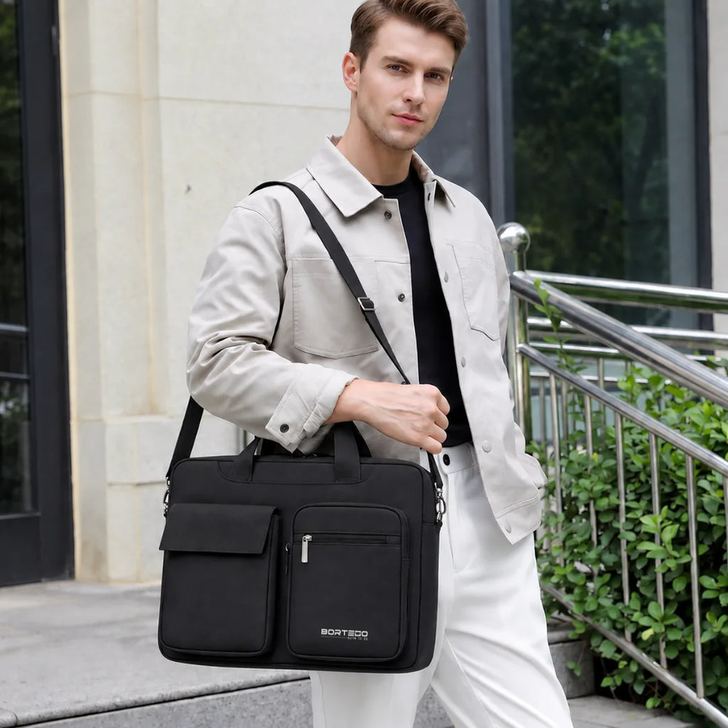A man wearing a light beige jacket, black shirt, and white pants carries a black BORTEGO business bag with a top handle and shoulder strap in an outdoor urban setting.