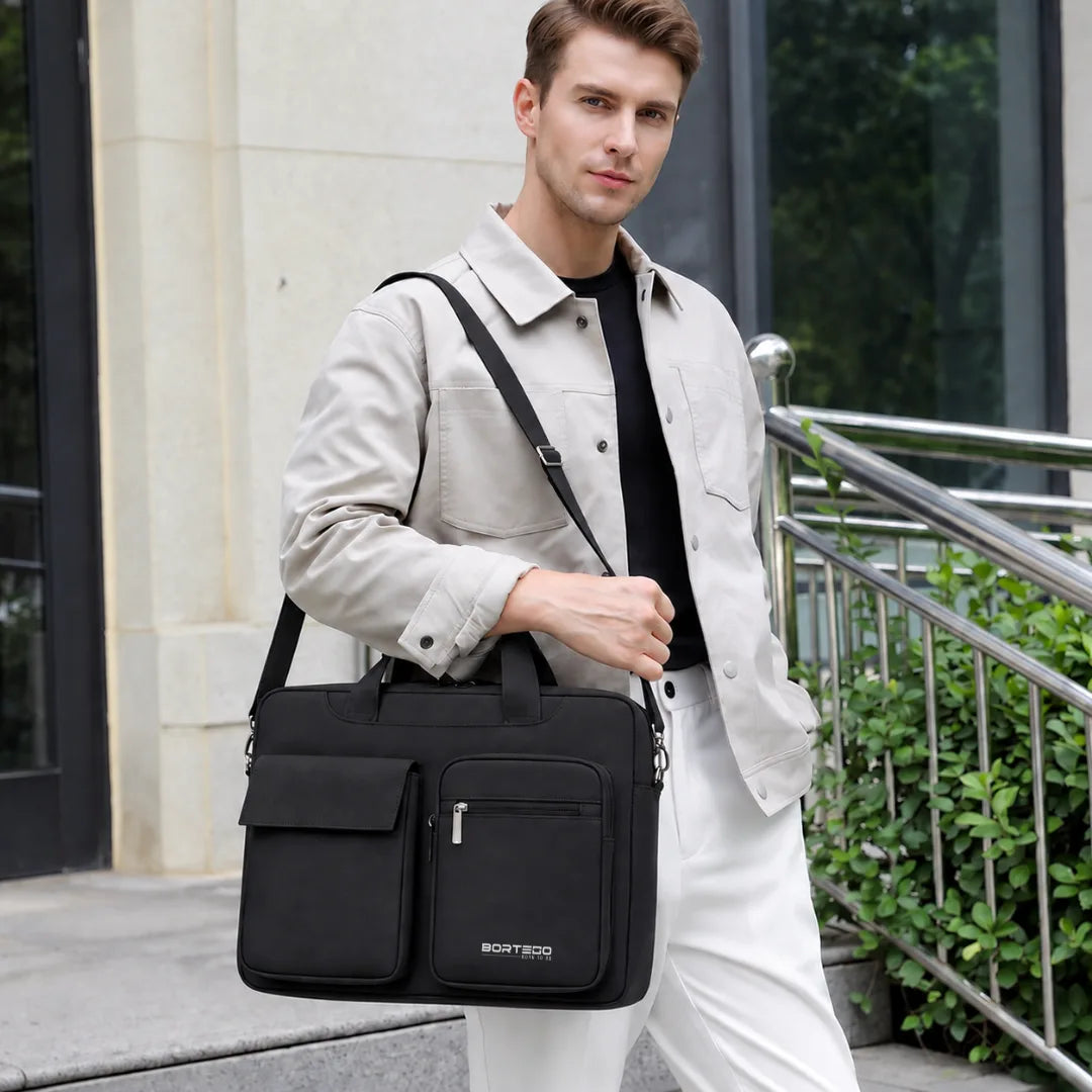 A man wearing a light beige jacket, black shirt, and white pants carries a black BORTEGO business bag with a top handle and shoulder strap in an outdoor urban setting.