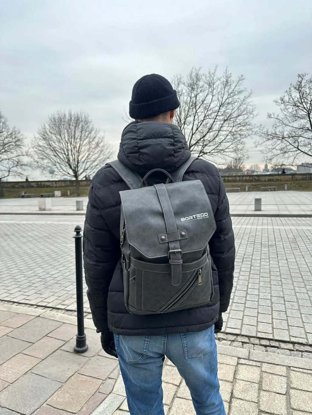 Person wearing a black jacket and blue jeans with a gray backpack walking on a paved path.