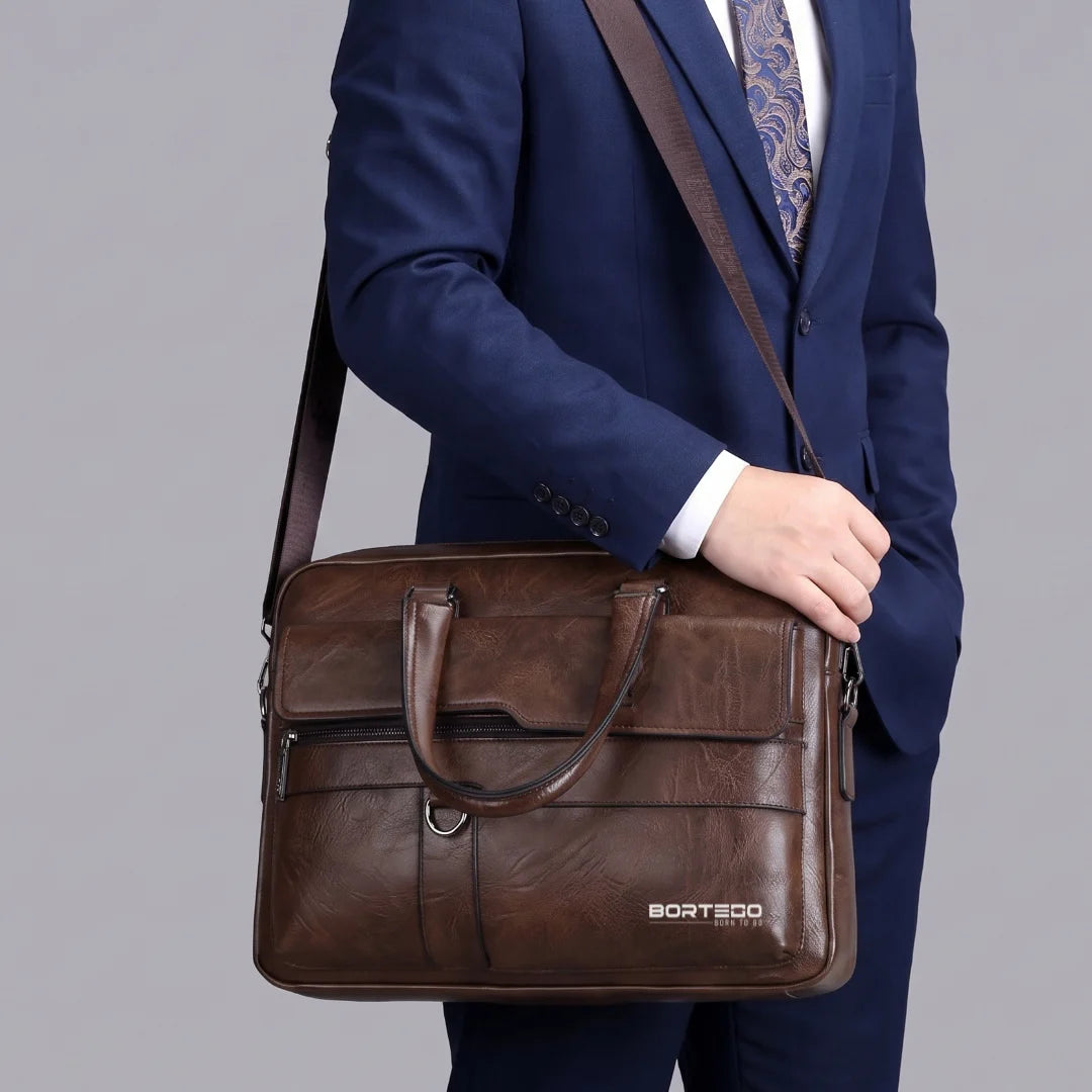 A man in a navy blue suit holding a brown leather briefcase-style shoulder bag with a long strap. The bag has top handles, a front flap pocket, and a small visible BORTEGO logo near the bottom right.