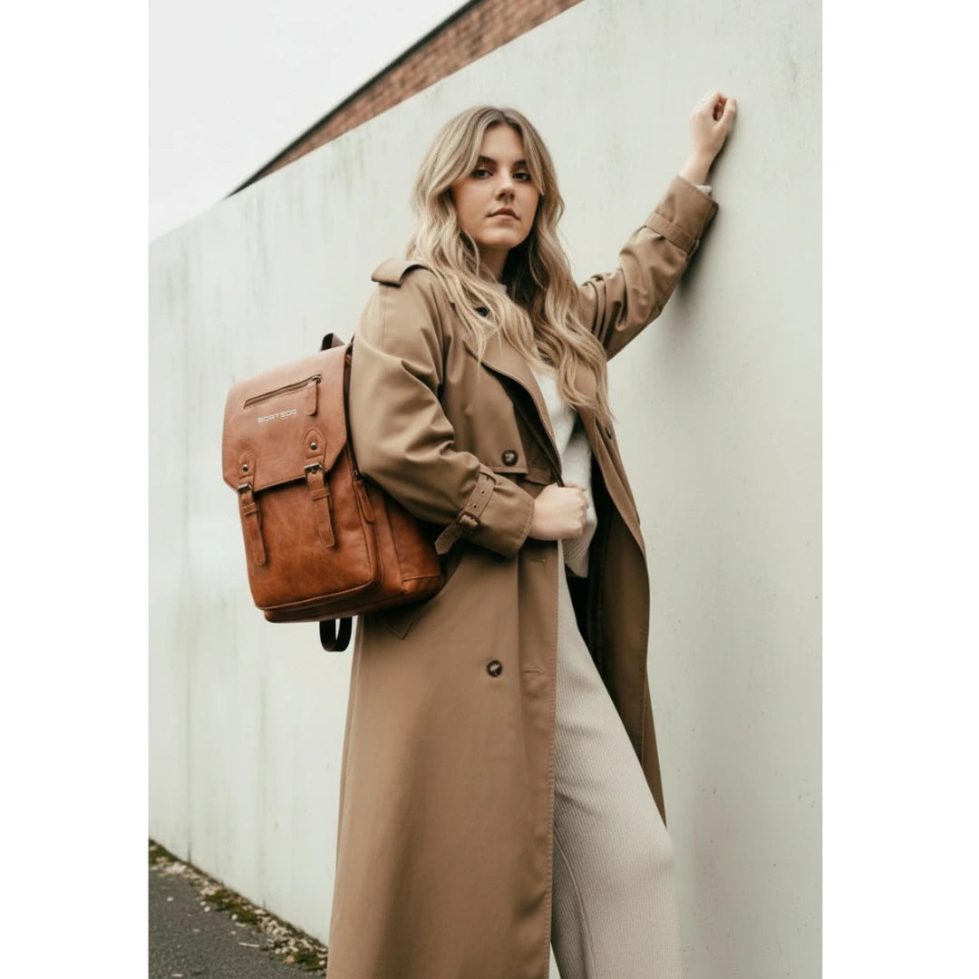 Woman in a long brown coat with a brown backpack against a white wall.