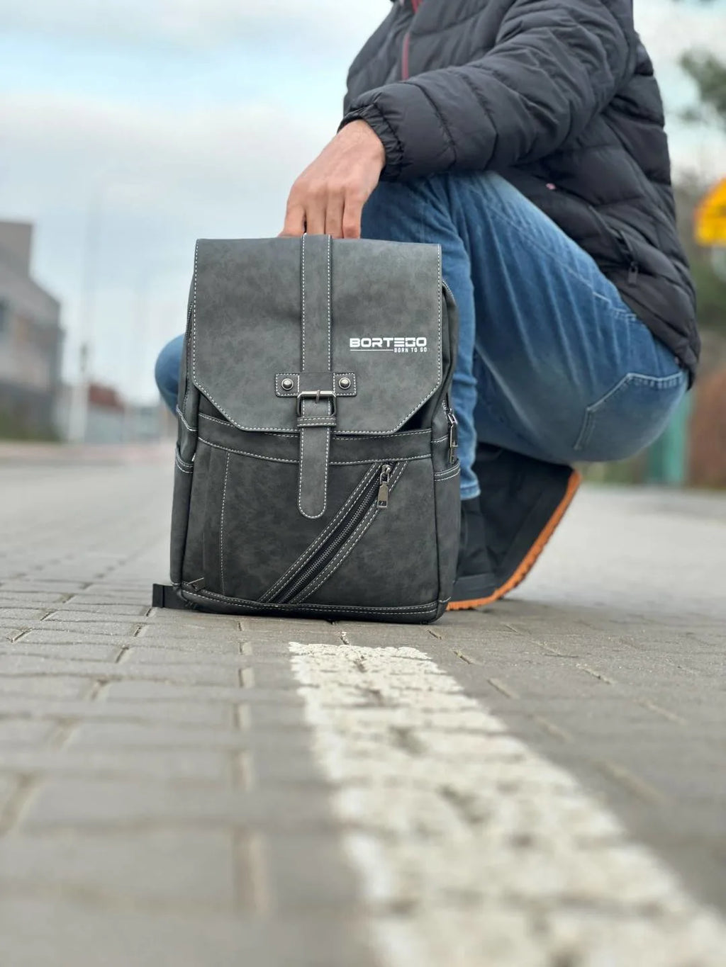 Person holding a gray backpack on a paved surface with blurred background