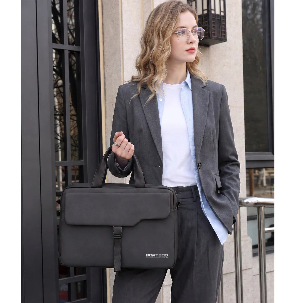 A woman wearing a gray blazer and white shirt holds a dark gray BORTSOGO briefcase-style shoulder bag in front of a building entrance.