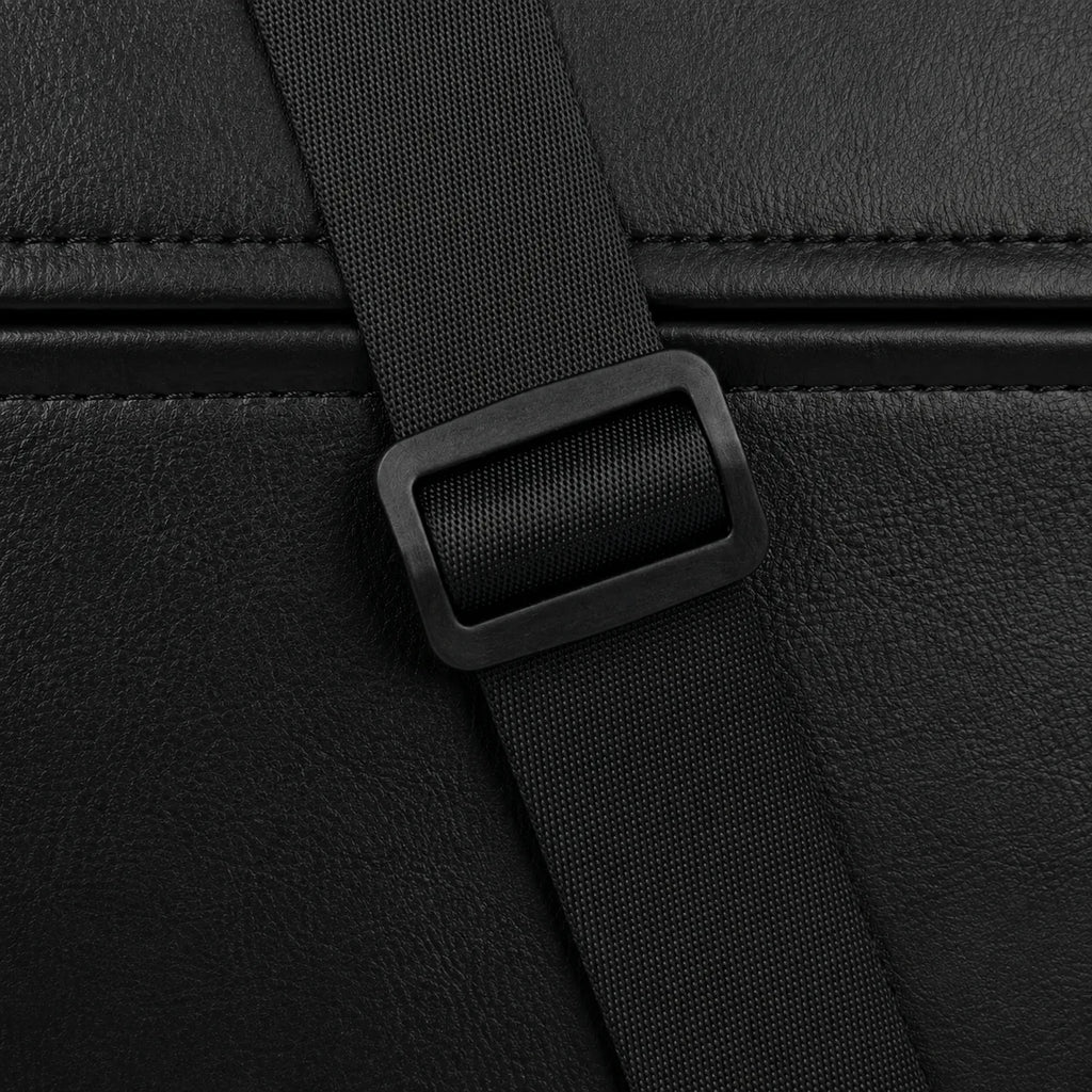 Close-up detail of a black textured leather or faux-leather bag with a matching black webbing strap and rectangular plastic buckle.