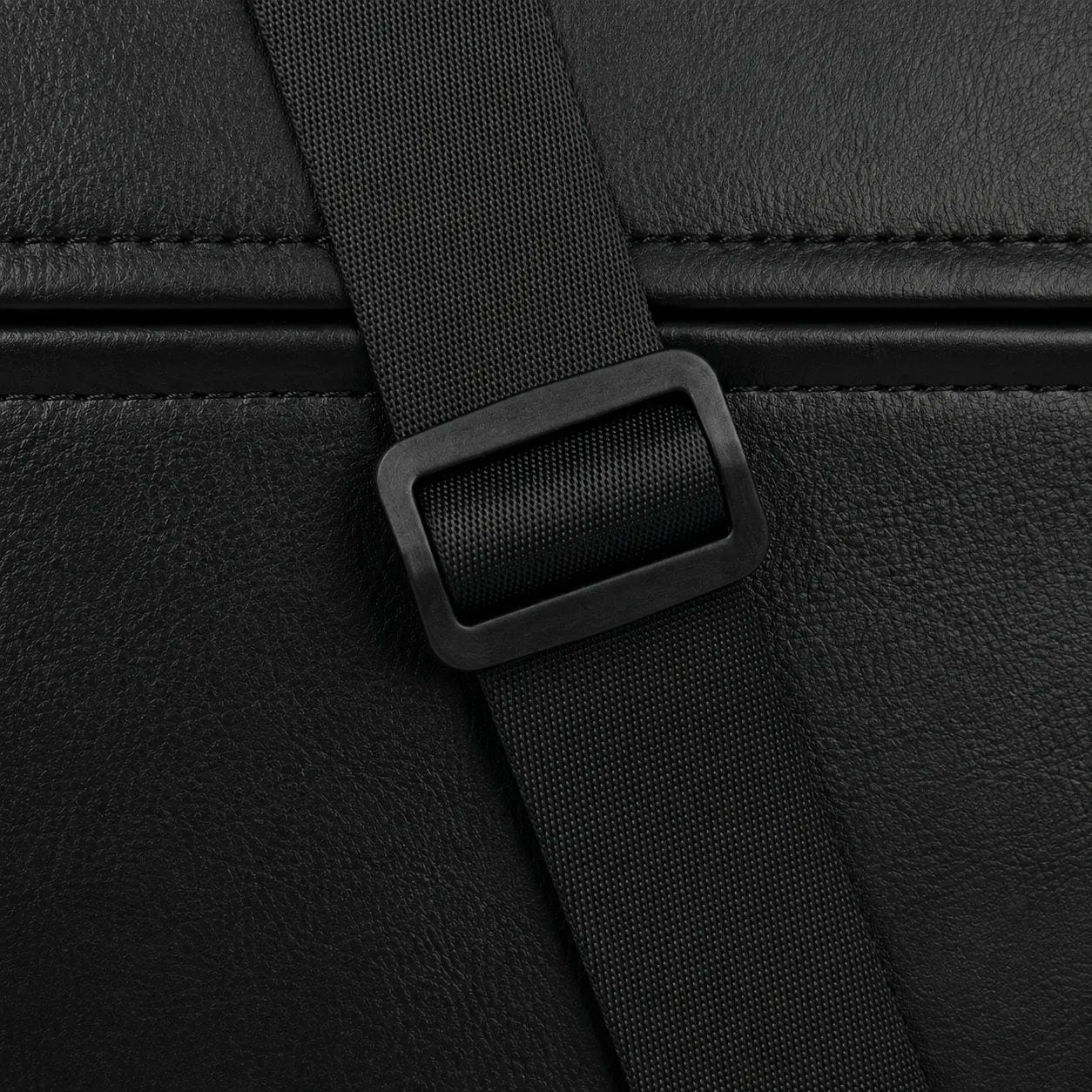 Close-up detail of a black textured leather or faux-leather bag with a matching black webbing strap and rectangular plastic buckle.