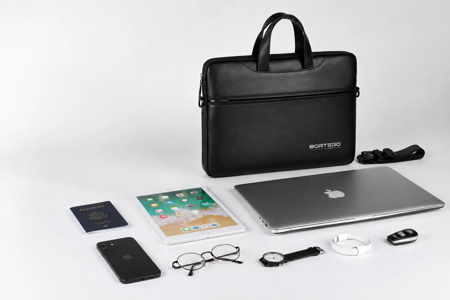 A black BORTEGO laptop briefcase standing upright on a light gray studio background, surrounded by lifestyle tech accessories including a closed silver laptop, tablet, smartphone, eyeglasses, watch, earbud case, and passport.
