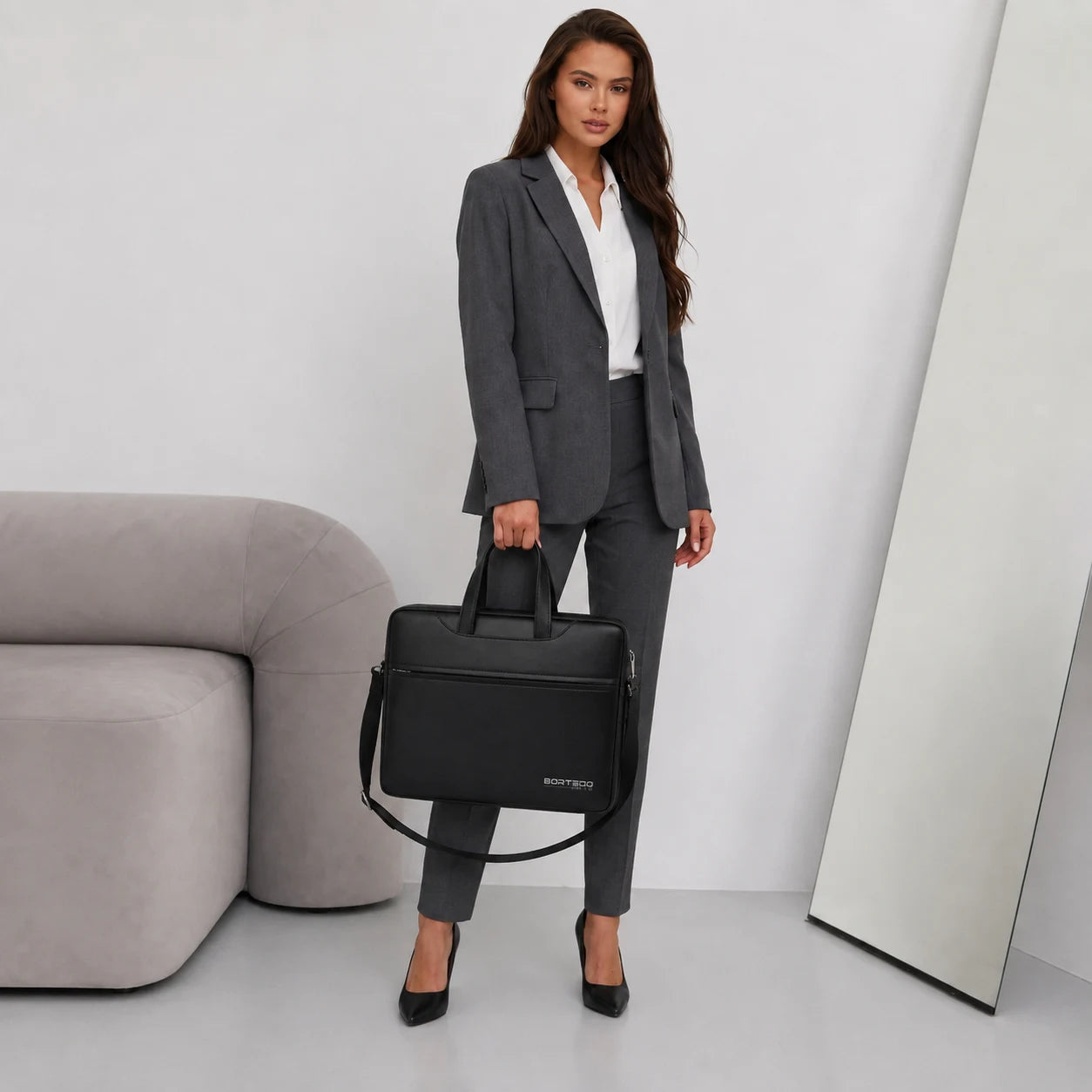 A woman in a gray blazer and trousers holds a black Bortegobriefcase in a minimalist studio setting with a gray sofa and a floor mirror.