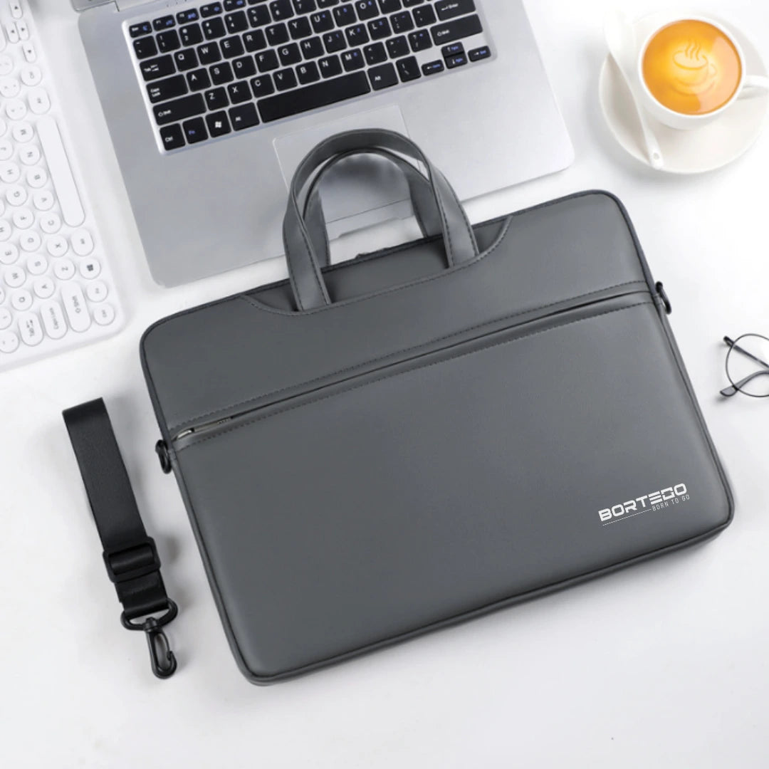 A dark gray BORTEGO laptop sleeve with top handles and a detachable shoulder strap, styled in a flat-lay on a white desk next to a laptop, keyboard, coffee cup, and glasses.