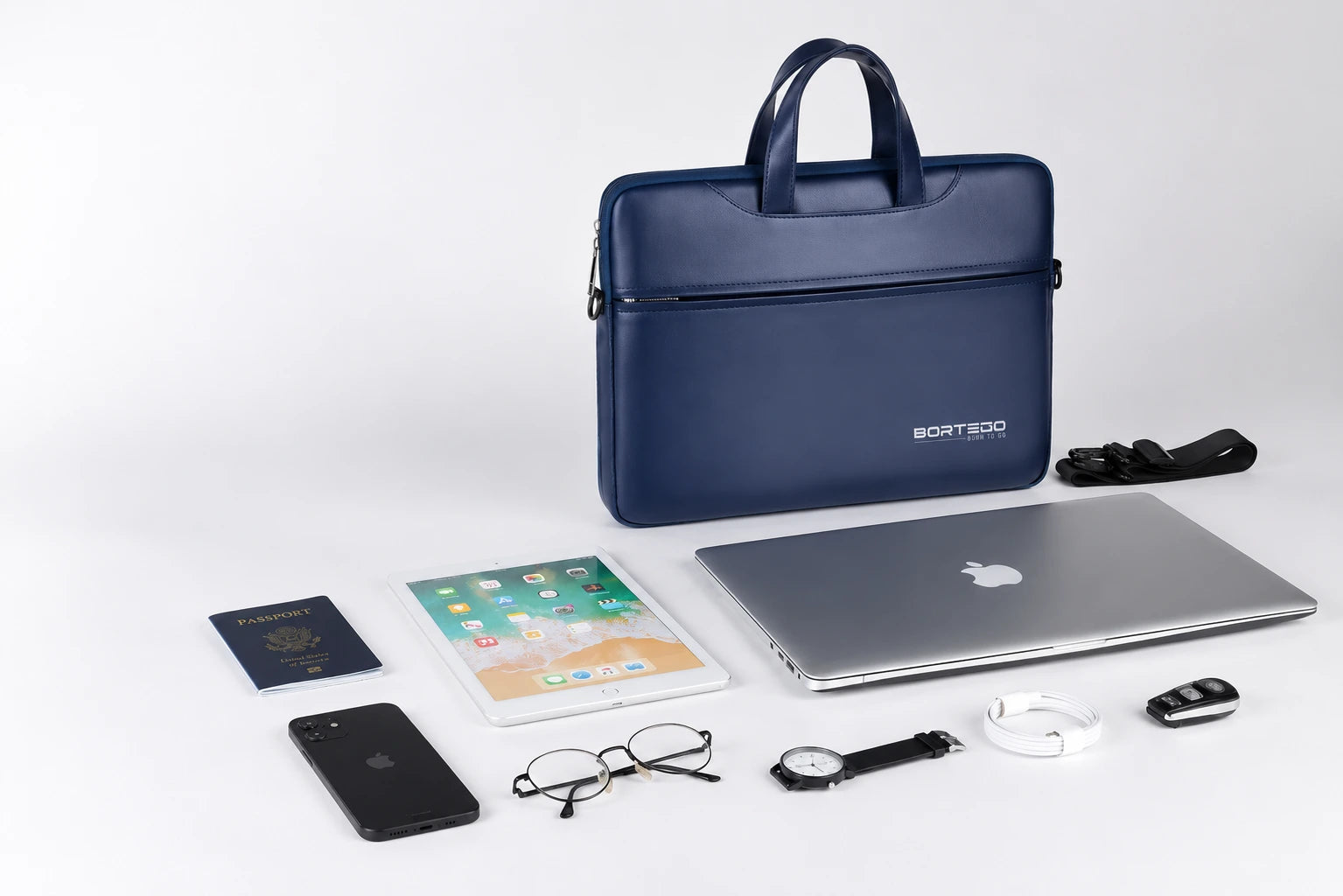 A navy blue laptop sleeve by BORTEGO is displayed on a clean white background, surrounded by various travel and office accessories including a silver laptop, tablet, smartphone, passport, glasses, watch, and key fob.