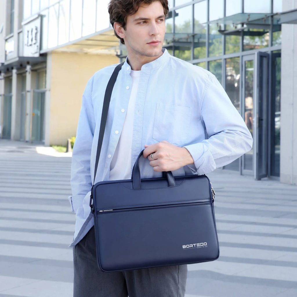 A man wearing a light blue button-down shirt and gray trousers carries a dark navy blue BORTEGO briefcase-style shoulder bag with top handles and a black strap, standing outdoors in front of a modern glass building.